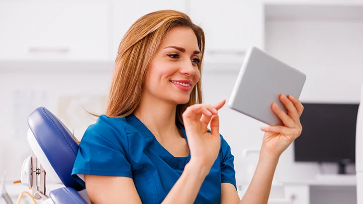 Mulher jovem sentada em cadeira de dentista sorrindo ao olhar tablet