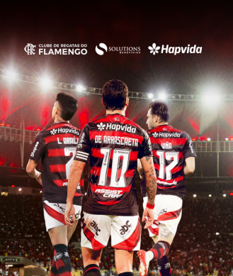 Jogadores do Flamengo com a marca Hapvida nos uniformes, diante de um estádio iluminado, com os logos do Flamengo, Solutions e Hapvida no topo da imagem.