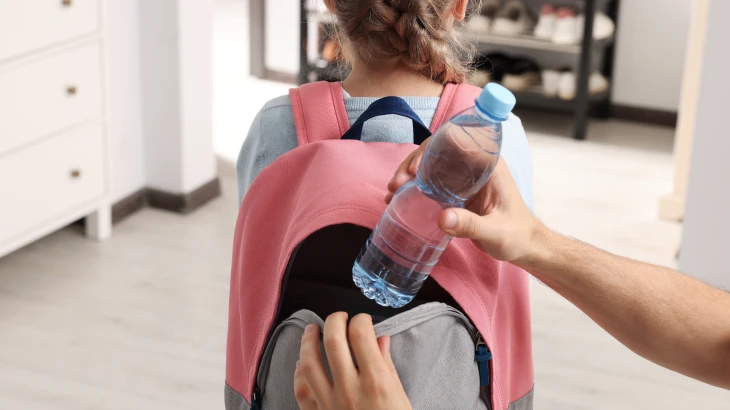Criança com uma mochila nas costa e uma mão colocando uma garrafa de água.