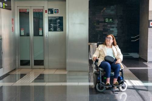 Mulher cadeirante aguarda em frente ao elevador com piso tátil em ambiente acessível e adaptado.